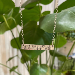 Tree necklace in silver bar style on chain shown hanging in front of a plant